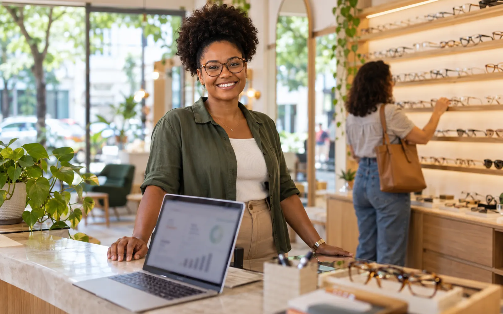 Dona de uma ótica sorrindo atrás do balcão com um notebook aberto, enquanto uma cliente escolhe armações ao fundo.