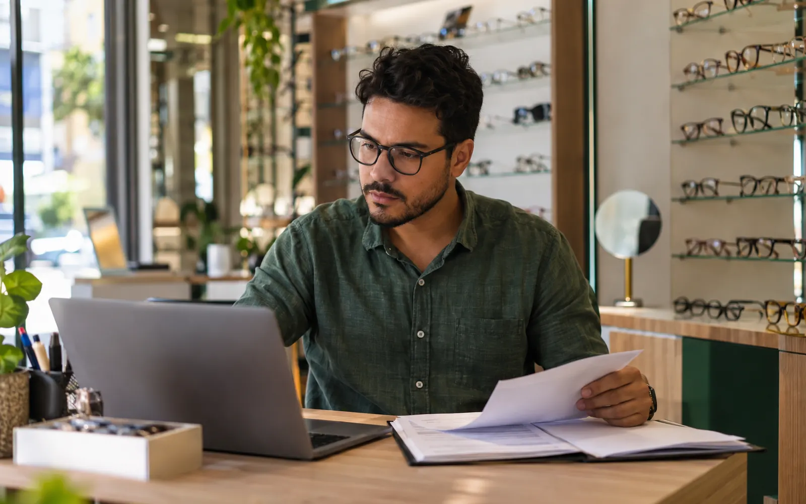 Pequeno empresário de ótica sentado a uma mesa com notebook e uma pasta de documentos em uma loja compacta.