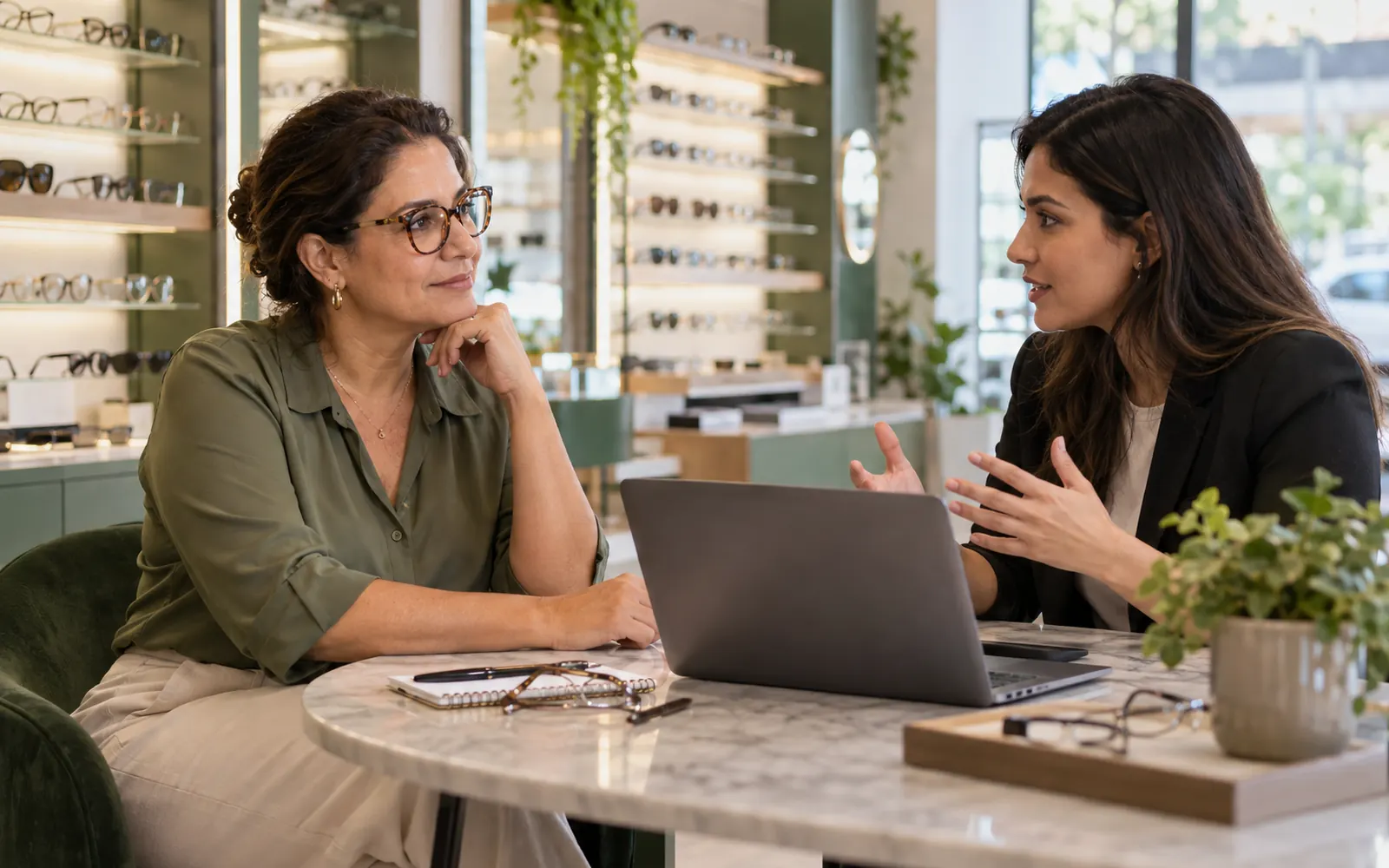 Reunião entre a dona de uma ótica e uma profissional de marketing com notebook, em uma mesa na loja.