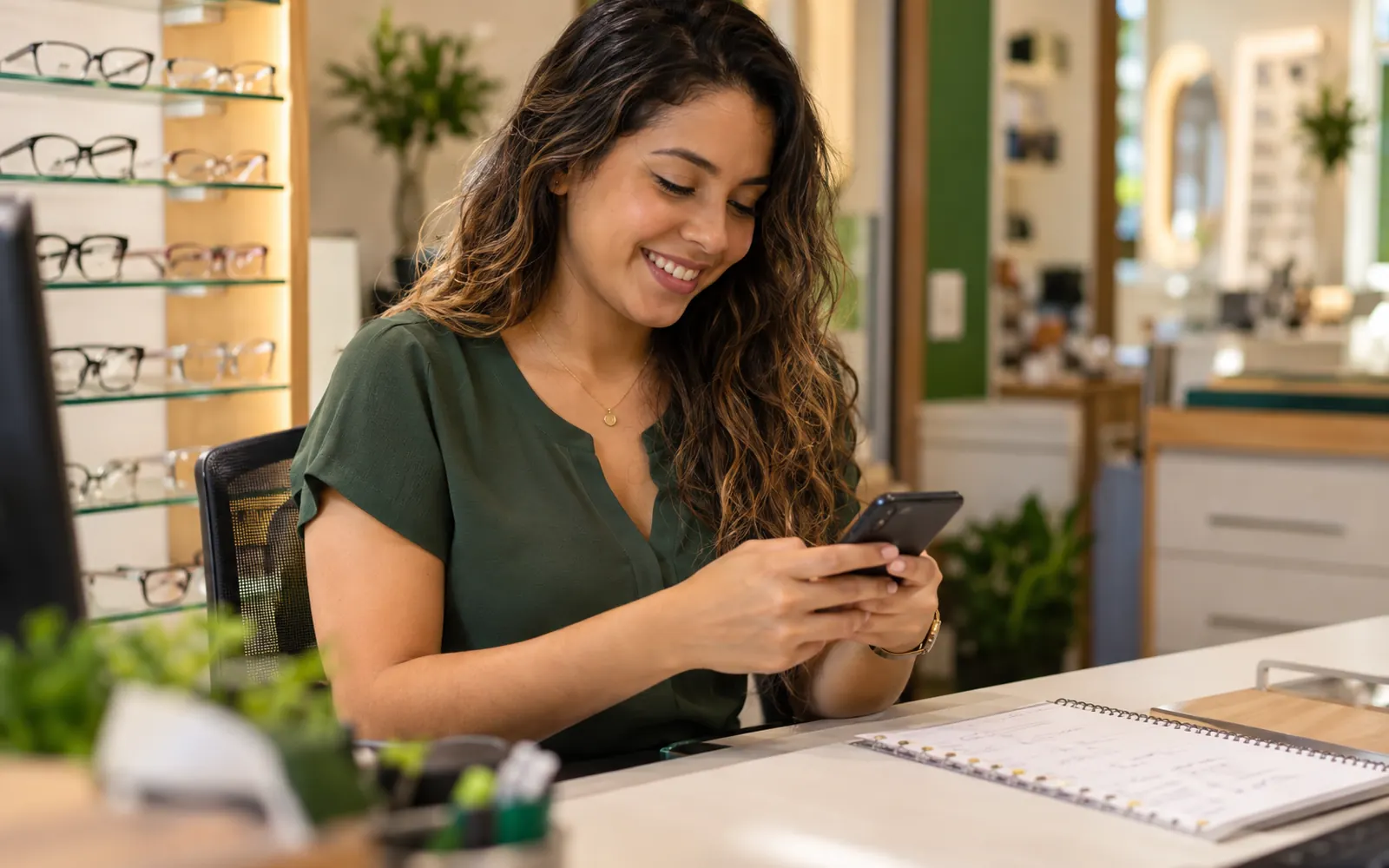 Atendente sorrindo ao digitar no celular em uma mesa de ótica, com um caderno de anotações ao lado.