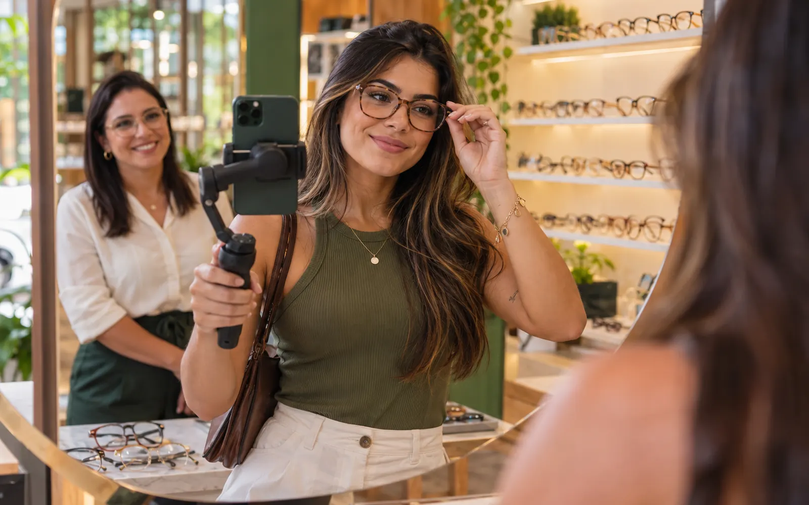 Influenciadora gravando um vídeo no celular dentro de uma ótica, experimentando armações diante do espelho.